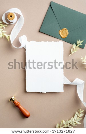 Ripped sheet of paper, silk ribbon, green envelope with wax seal, dried flowers on pastel beige background. Wedding invitation card template. Flat lay, top view, copy space.