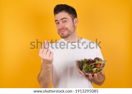 Young handsome Caucasian man holding a salad bowl against yellow background, making money gesture.
