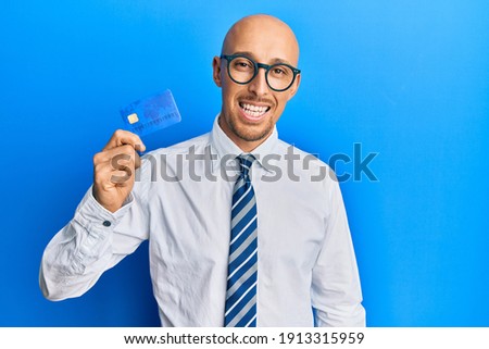 Bald man with beard holding credit card looking positive and happy standing and smiling with a confident smile showing teeth 