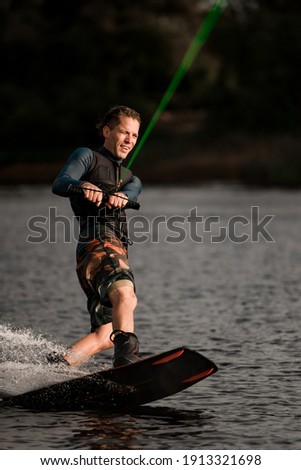 adult cheerful man in wetsuit holds taut bright cable in his hands and balancing on splashing wave