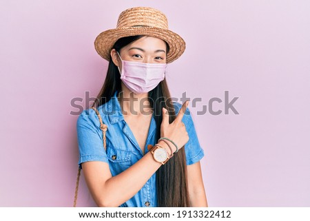 Young chinese woman wearing medical mask on summer smiling cheerful pointing with hand and finger up to the side 