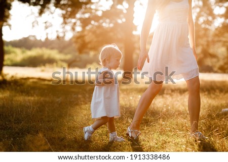 young beautiful mom is walking with little daughter in the sunny park . The concept of summer holiday outdoors. Mother's, baby's day. Spending time together. Sun light Royalty-Free Stock Photo #1913338486