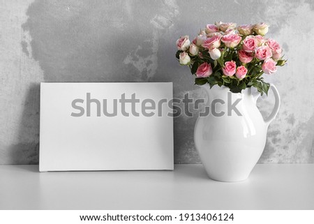 Blank canvas mockup on white table with pink flowers in vase on concrete wall background. White stretched canvas, interior decor