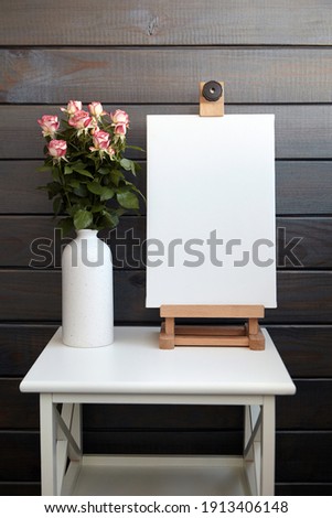 Blank canvas mockup on easel on white table with roses in vase on brown wall background. White stretched canvas, interior decor