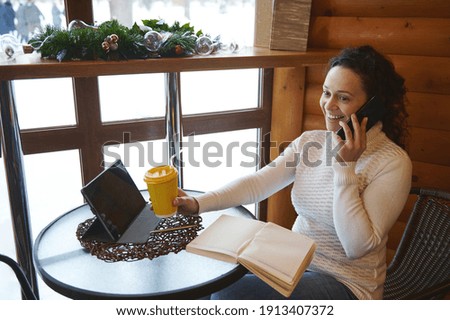 Smiling woman holding coffee paper cup and talking by mobile phone while sitting at cafeteria.