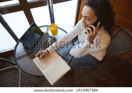 Top view of a negotiating woman by phone and writing on a diary while sitting near window at a wooden cafe