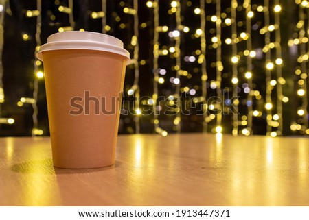 Disposable paper cup on table on wooden table against glowing lights background. New year and Christmas festive mood concept. Mockup for advertising, design with copy space 