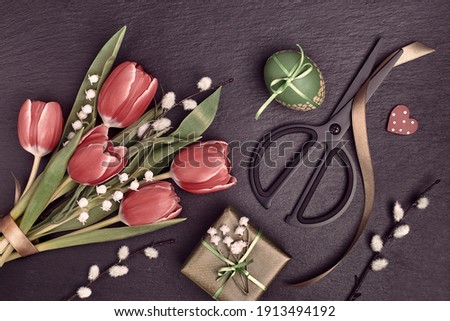 Overhead view, flat lay. Arrangement of Easter, springtime decorations. Wrapped gifts, red tulips, pussy willow, lily of the valley flowers. Painted Easter eggs, scissors on black slate background