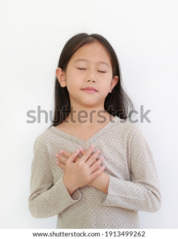 Portrait of Asian little girl closed eyes and holding hands on heart gesture of love. Kid place arms on chest isolated on white background.