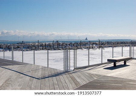 An angled metal fence casts a shadow on a wood deck with bench on the shore of a frozen Lake Michigan in Milwaukee Wisconsin