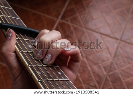 A woman with glitter nail polish plays an A minor chord on an accoustic guitar, fitted with a capo, February 2021 during COVID-19 lockdowns. Royalty-Free Stock Photo #1913508541