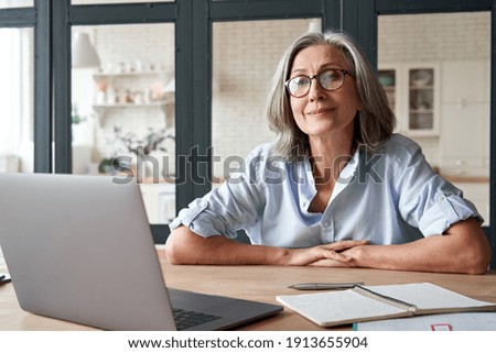Smiling stylish mature middle aged woman sits at desk with laptop, portrait. Happy older senior businesswoman, 60s grey-haired lady wearing glasses looking at camera sitting at office table. Headshot. Royalty-Free Stock Photo #1913655904