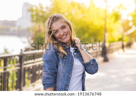 Portrait of a happy and attractive blonde Caucasian young woman in a casual denim jacket outdoors in a park on a sunny autumn day. A concept of lifestyle, happiness and joy. Royalty-Free Stock Photo #1913676748