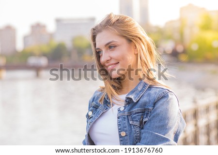Portrait of a happy and attractive blonde Caucasian young woman in a casual denim jacket outdoors in a park on a sunny autumn day. A concept of lifestyle, happiness and joy. Royalty-Free Stock Photo #1913676760