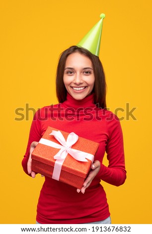 Cheerful female in red turtleneck and green party hat smiling for camera and demonstrating present during birthday celebration against yellow background