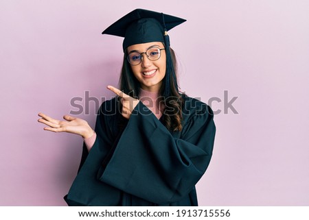 Young hispanic woman wearing graduation cap and ceremony robe amazed and smiling to the camera while presenting with hand and pointing with finger.  Royalty-Free Stock Photo #1913715556