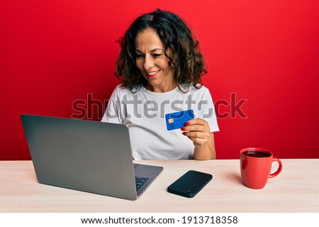 Beautiful middle age woman working at the office using laptop and credit card looking positive and happy standing and smiling with a confident smile showing teeth 