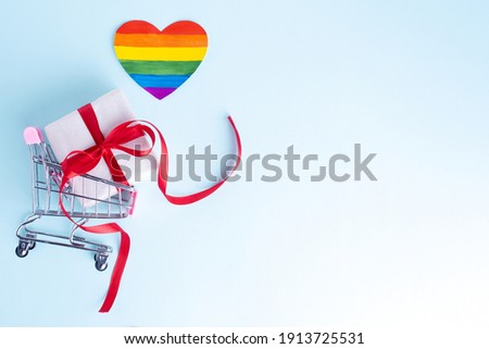 Gift box with a red ribbon in a copy of a shopping cart and a paper rainbow heart shape on a gentle blue background. Online shopping concept. online congratulations concept. LGBT concept