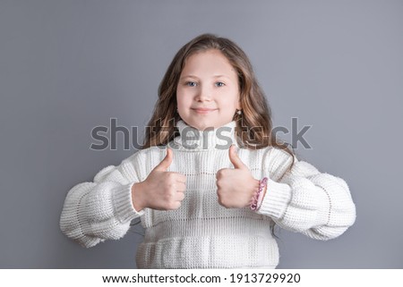Portrait of a young attractive little girl with blond long flowing hair in a white sweater smiling shows thumbs up,like sing on a gray studio background. Place for text. Copy space