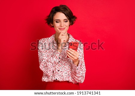 Photo portrait of curious woman touching face chin with finger holding phone in one hand isolated on bright red colored background