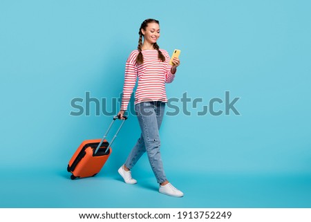 Full length photo of cute pretty young woman dressed striped shirt walking holding luggage modern device isolated blue color background