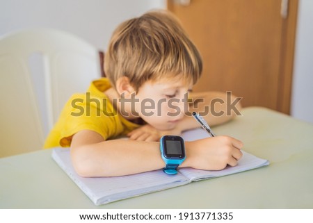 Little boy sitting at the table and looking smart watch. Smart watch for baby safety. The child makes school lessons, listening to music, calling friends