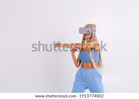 Studio photo of a young attractive woman in a warm blue fashionable suit wearing virtual reality glasses on a white background plays a boxing fight