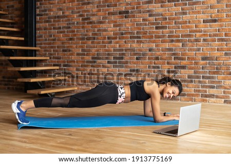 Attractive and smiling african-american woman in sportswear warm up, doing plank exercises near laptop at home, watching video training
