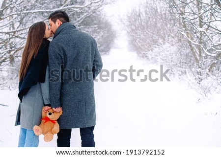 Behind photo of caucasian couple walking in the winter snowy park. Family sweet kiss, holding together a brown plush teddy bear. Infinity white natural background with empty space for advertisement