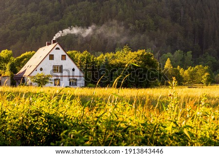 Rural view, a traditional polish country cottage house with smoke from the chimney next to pine forest. Rural cottage in the distance at sunset Royalty-Free Stock Photo #1913843446