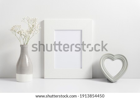 Valentine's Day background. White photo frame, vase with dried flowers, heart on white table. Front view. Place for text, copy space, mockup