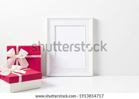 White blank photo frame, two pink gifts with white satin bow on white table. Front view. Place for text, copy space, mockup