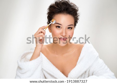 Beautiful woman using derma roller for her facial skin. Photo of asian woman after shower on white background. Beauty and skin care concept