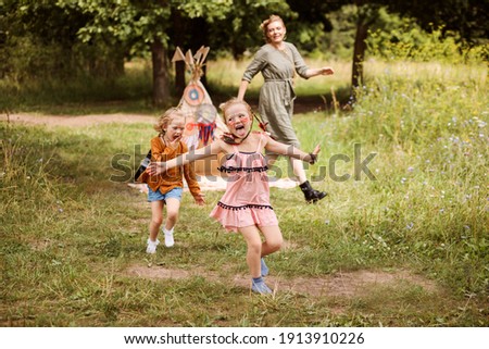 Children are playing outdoors with their mom. Family is dressed in boho style, sisters have native american's makeup on faces. Wigwam in the background. Happy childhood, summer holidays. 