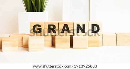 Wooden cubes with letters on a white table. The word is GRAND. White background with photo frame, house plant.