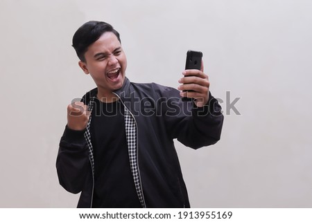 Portrait of happy young Asian man in black jacket holding mobile phone shaking fits and celebrating success. Winner of competition concept. Isolated image on gray background