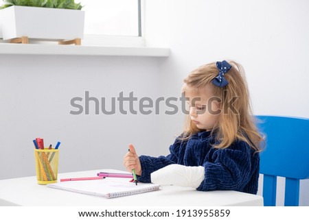 Little girl with hand in cast sitting at table trying draw with markers.