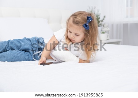 Little girl with hand in cast laying in bed using smartphone, watching cartoon or education video.