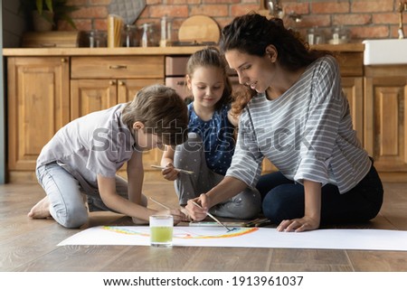 Drawing colorful dream. Happy young mom rest sit on warm floor paint big fantasy picture with small daughter son. Inspired millennial female tutor nurse and two junior children pupils use watercolors