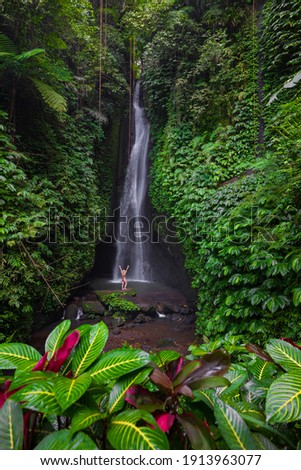 Travel lifestyle. Young traveler woman wearing bikini at waterfall in tropical forest. Excited Caucasian woman raising arms in front of waterfall. View from back. Leke Leke waterfall, Bali.