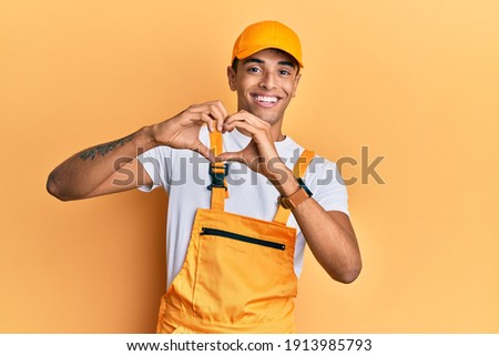 Young handsome african american man wearing handyman uniform over yellow background smiling in love doing heart symbol shape with hands. romantic concept. 