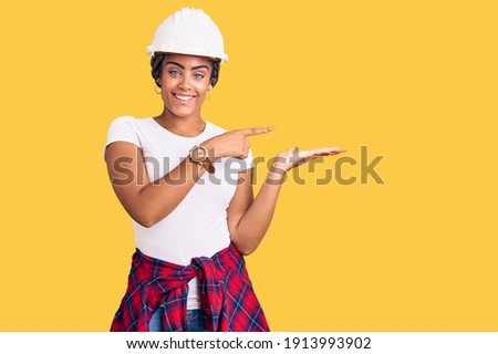 Young african american woman with braids wearing hardhat and builder clothes amazed and smiling to the camera while presenting with hand and pointing with finger. 