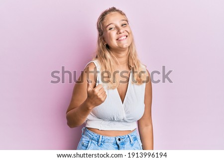 Young blonde girl wearing casual clothes doing happy thumbs up gesture with hand. approving expression looking at the camera showing success. 