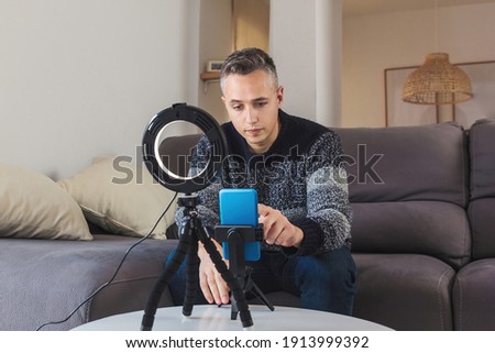 Stock photo of Young man millennial influencer sit on sofa preparing a setup with mobile phone and lighting for vlogging or video call. Social media video blog recording concept