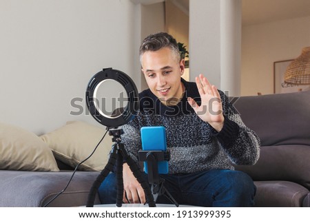 Stock photo of Young man millennial influencer sit on couch is vlogging, recording and creating online content with smartphone and lights. Social media video channel recording concept