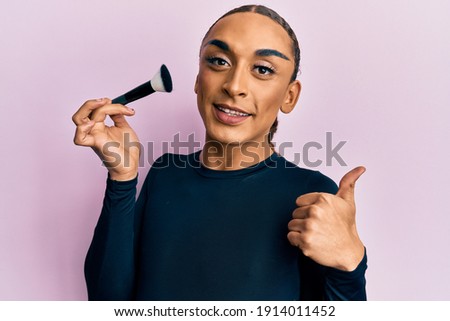 Hispanic man wearing make up and long hair holding makeup brush smiling happy and positive, thumb up doing excellent and approval sign 