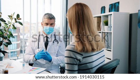 Rear of Caucasian beautiful blonde female patient sitting in hospital cabinet listening to middle-aged doctor in medical mask, general practitioner showing vaccines to woman on consultation virus cure Royalty-Free Stock Photo #1914030115