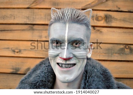 A young guy in cat makeup, with cat lenses and ears looks and smiles at the camera on a wooden background in a gray fur coat.