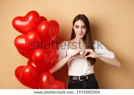 Image of beautiful young woman smiling and showing heart gesture, I love you sign, standing near romantic balloons on Valentines day, beige background