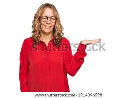 Middle age blonde woman wearing casual shirt over red background smiling cheerful presenting and pointing with palm of hand looking at the camera. 